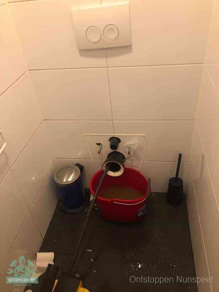 Een ontstopte wand-wc met verwijderde toiletpot waar een ontstoppingsveer in de wanduitgang verdwijnt, het achterhalen waar de verstopping zit in Nunspeet
