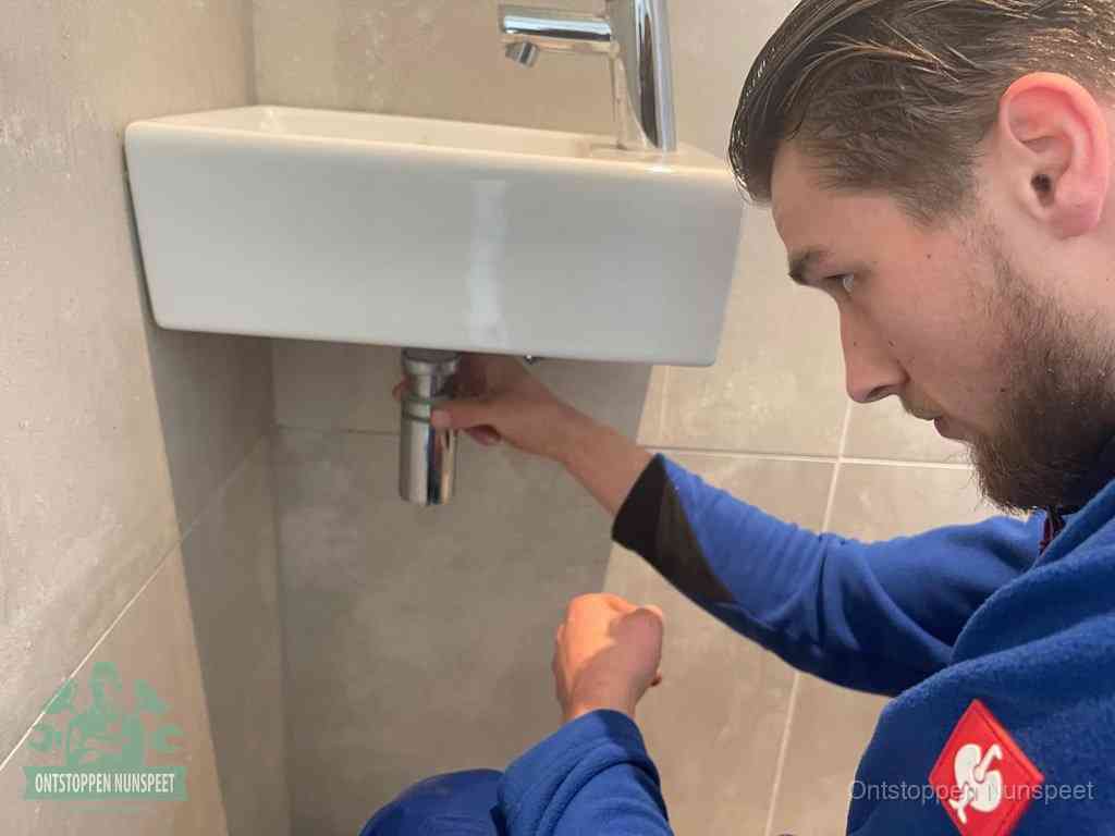 Een monteur draait met de hand de zwanenhalssifon onder een fonteinwasbak in de toiletruimte los om de bron van rioollucht in Nunspeet te vinden