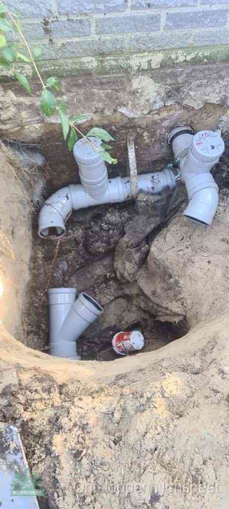 Een opgegraven bouwput langs een huismuur met grijze PVC rioolbuizen die op het hoofdriool worden aangesloten, typisch leidingwerk per wijk in Nunspeet